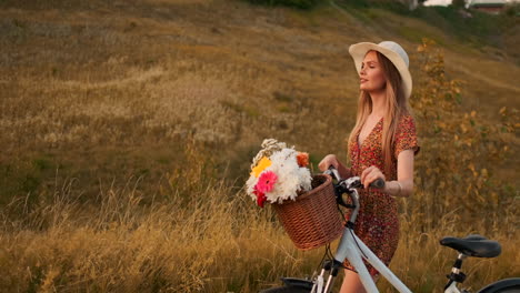 Slow-motion:-Young-sexy-smiling-blonde-woman-in-hat-and-light-brown-dress-walking-with-bike-and-flowers-in-basket-on-field-in-summer-at-sunset.-Moves-in-the-direction-of-the-camera.